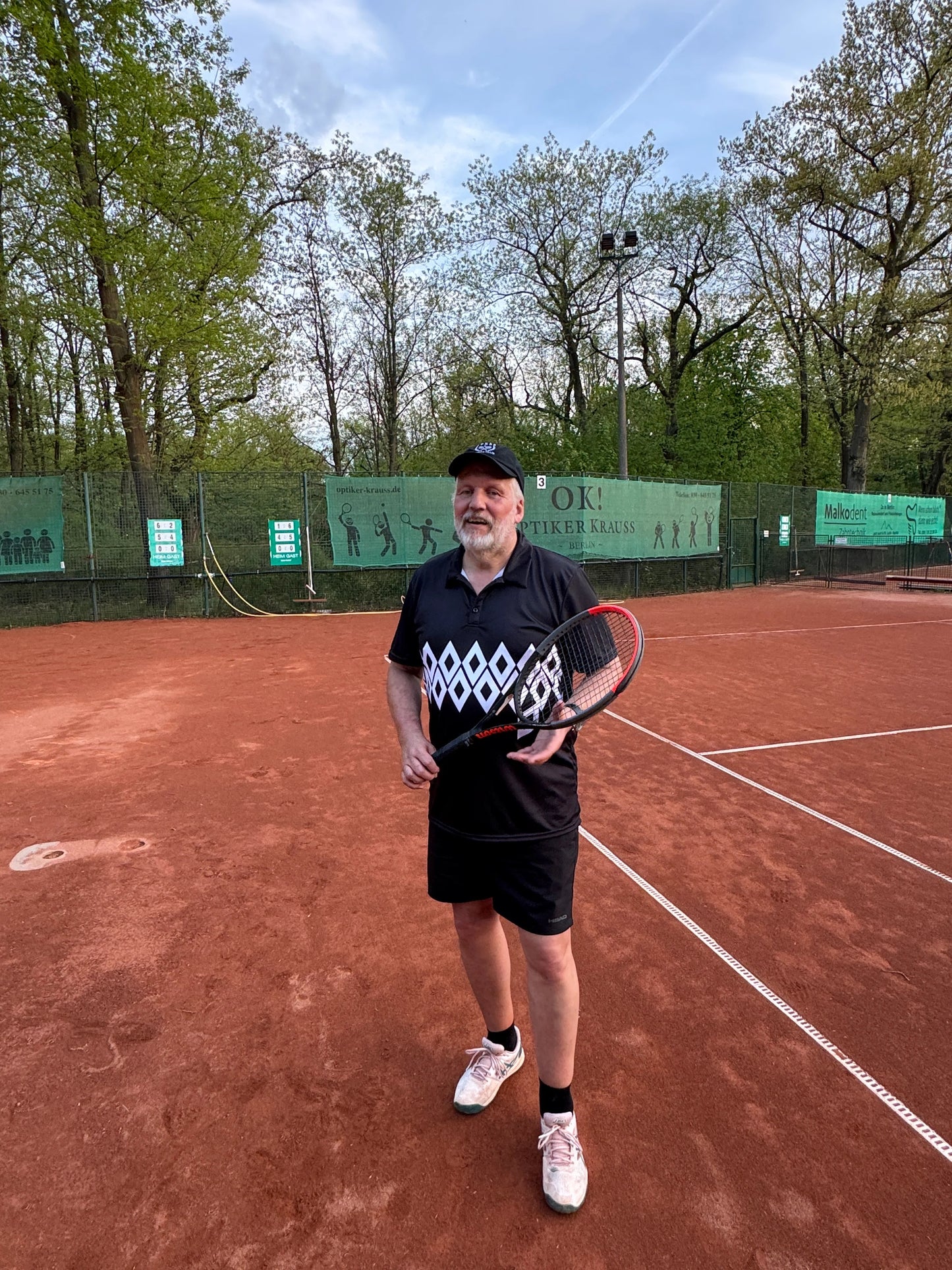 Laluedesign founder in diamond shape shirt with Tennis racket on a clay court with trees and blue sky in the background