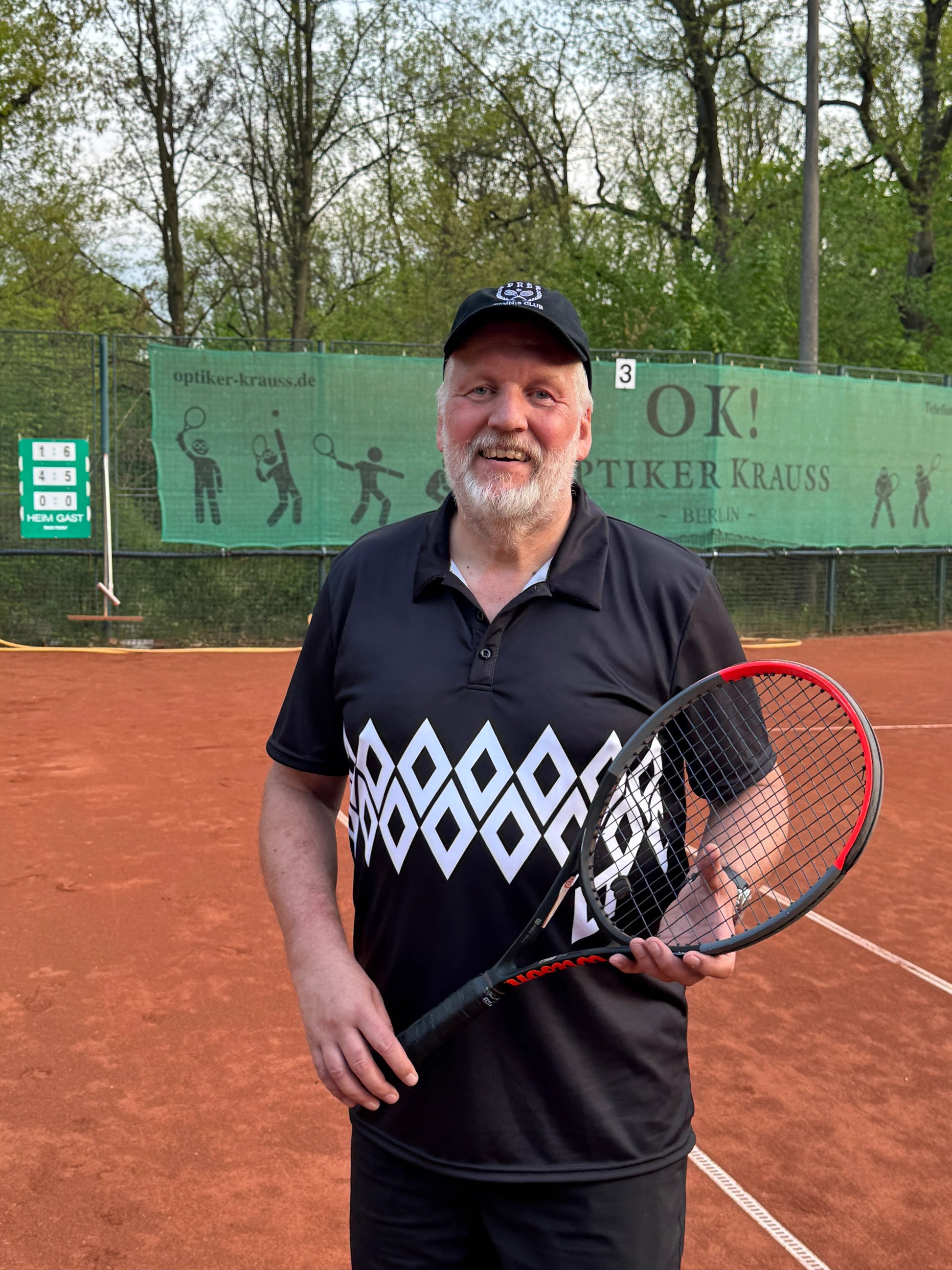 Laluedesign founder in diamond shape shirt with Tennis racket on a clay court with trees and blue sky in the background