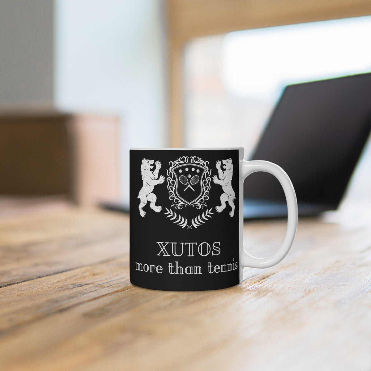 A black ceramic mug with a white handle and a white printed tennis-themed crest and the text 'XUTOS more than tennis', sitting on a wooden table with a blurred laptop in the background.