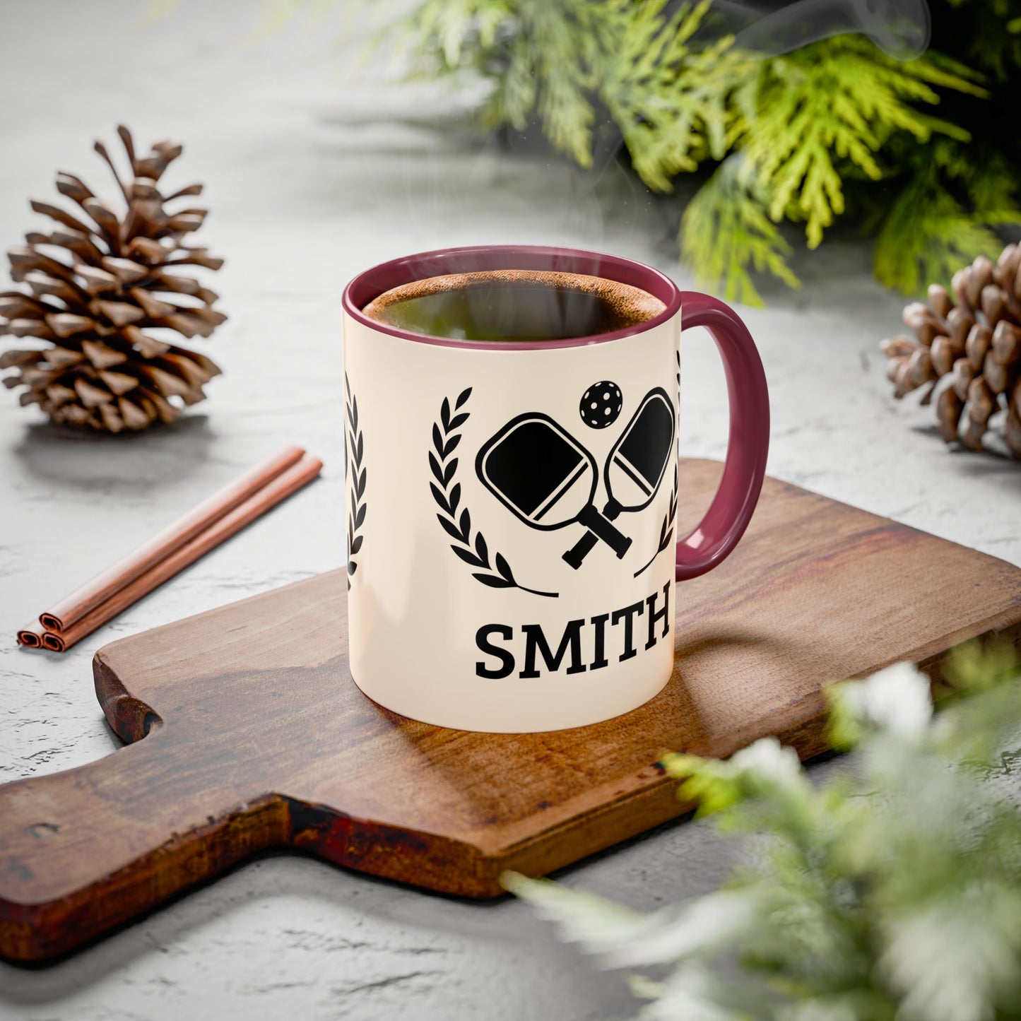 pickleball mug A personalized ceramic mug with a cream body, dark red handle and rim, and a black ping-pong graphic with the name 'SMITH' printed below. The mug sits on a wooden cutting board surrounded by pinecones, cinnamon sticks, and green foliage on a gray stone surface.