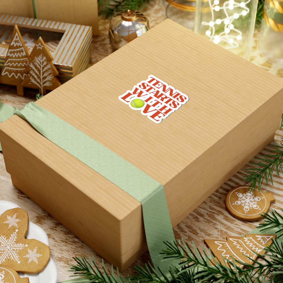 Gift box with a green ribbon and decorative text on a festive background with cookies and lights.