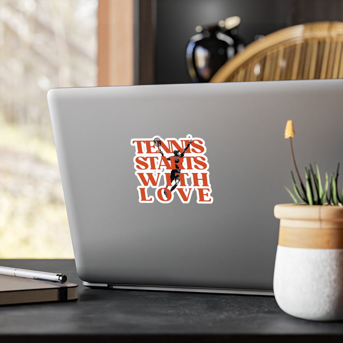Laptop with a 'Tennis Starts with Love' sticker on a desk with a plant and pen.