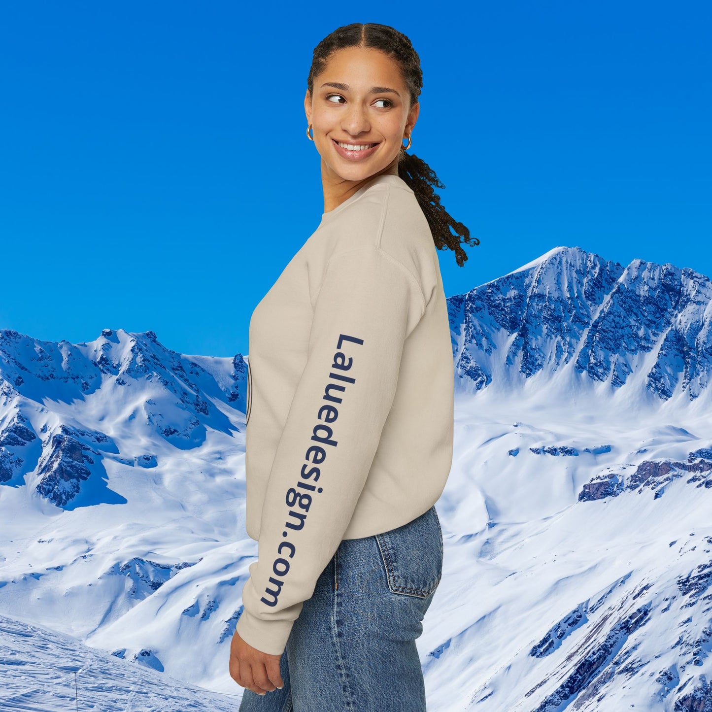 Après Ski Sweatshirt A woman wearing a beige crewneck sweatshirt with 'Laliedesign.com' printed vertically in navy on the left sleeve, posed against a digital background of snowy mountains and a blue sky.