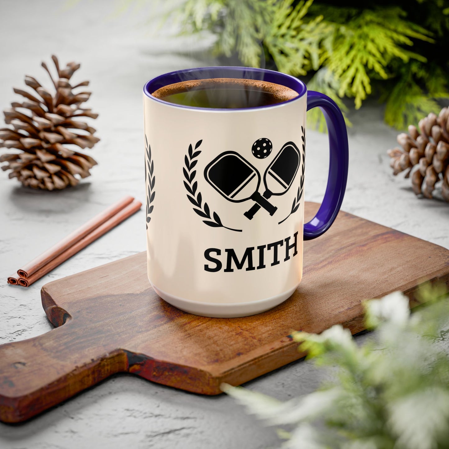 pickleball mug A cream-colored ceramic mug with a dark blue handle and rim, featuring a black graphic of crossed ping-pong paddles and the name 'SMITH' printed below. The mug is filled with a hot beverage and sits on a wooden cutting board surrounded by pinecones and greenery.