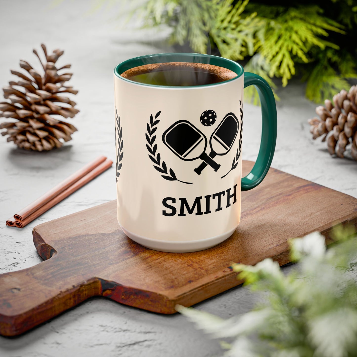 pickleball mug A cream-colored ceramic mug with a dark blue handle and rim, featuring a black graphic of crossed ping-pong paddles and the name 'SMITH' printed below. The mug is filled with a hot beverage and sits on a wooden cutting board surrounded by pinecones and greenery.
