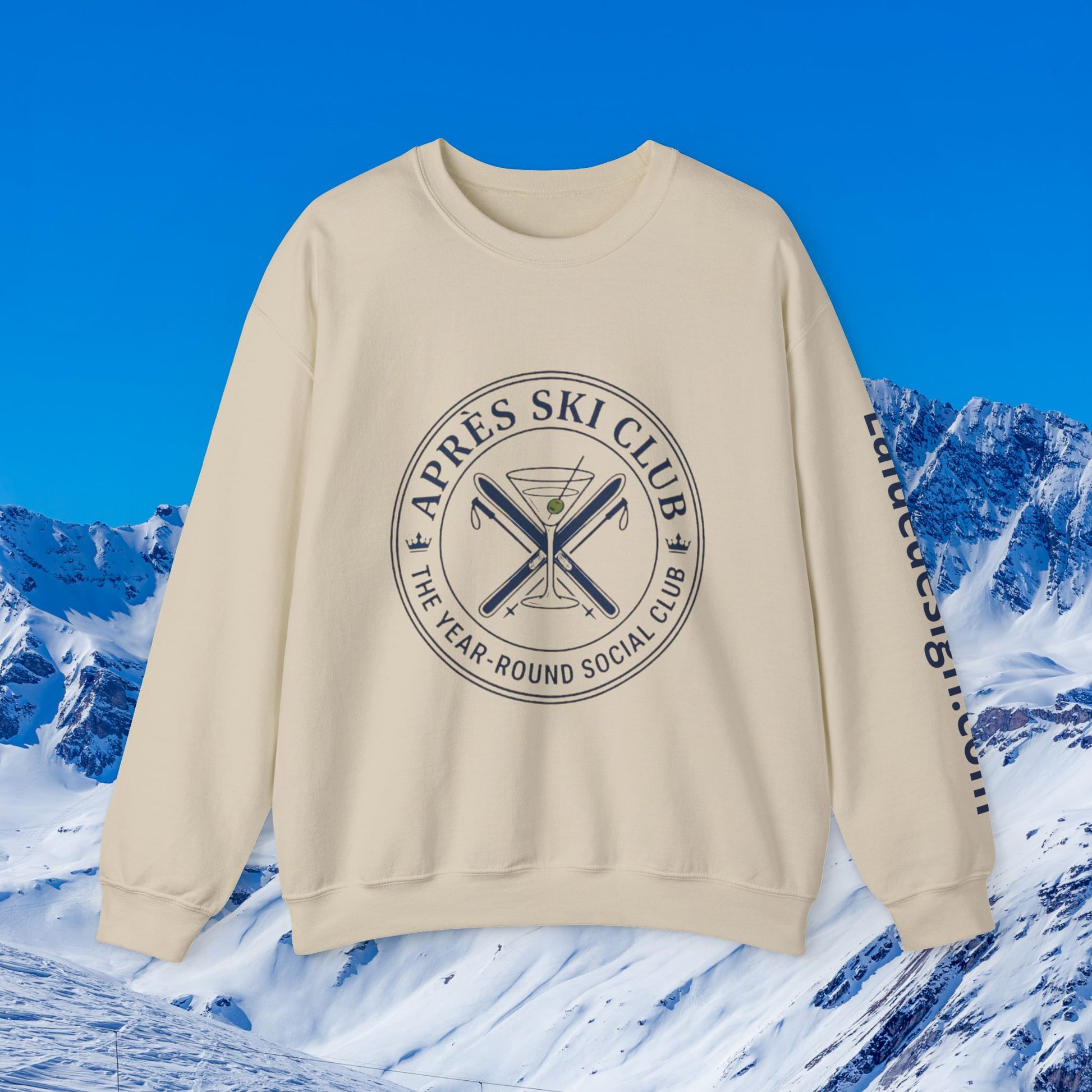 Après Ski Sweatshirt A cream-colored crewneck sweatshirt featuring a circular graphic on the chest that reads 'APRÈS SKI CLUB THE YEAR-ROUND SOCIAL CLUB' around crossed skis and a martini glass, with 'After Ski Club' printed down the sleeve. The sweatshirt is displayed against a digital background of snowy mountains and a bright blue sky.