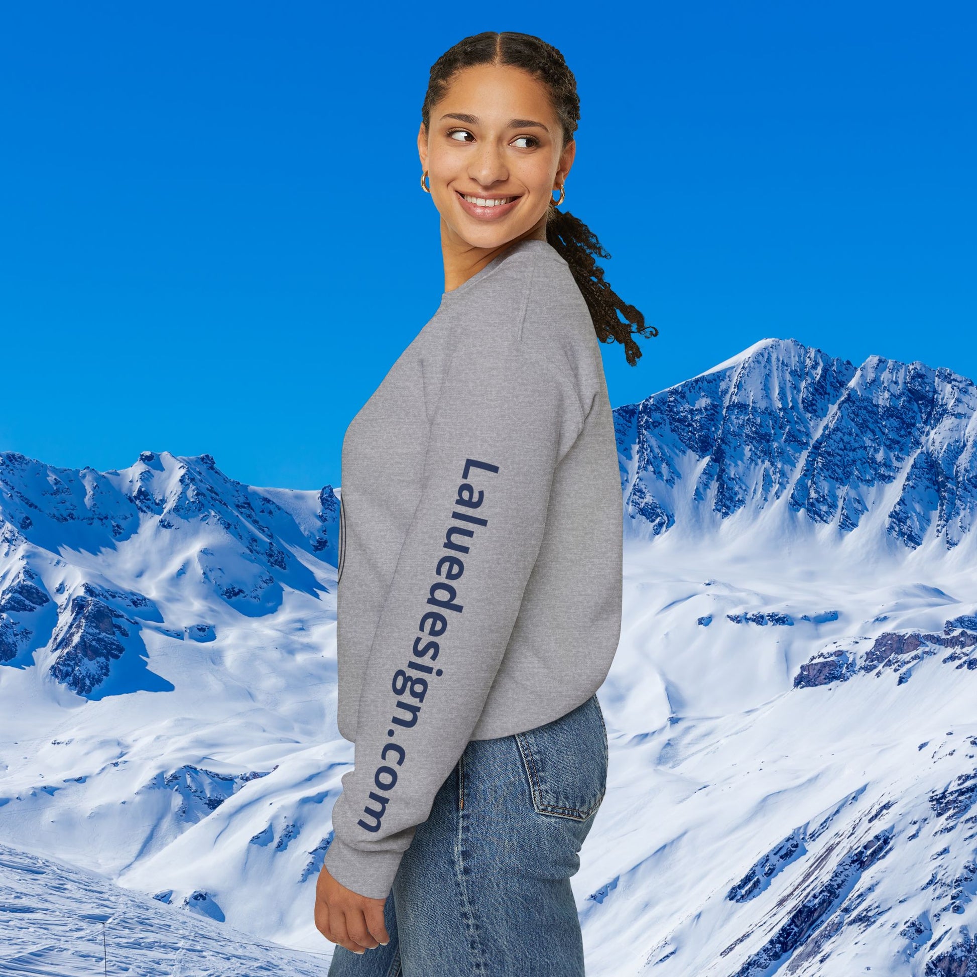 Après Ski Sweatshirt A woman wearing a light gray crewneck sweatshirt with 'Laliedesign.com' printed down the sleeve, set against a bright blue sky and snowy mountain background.