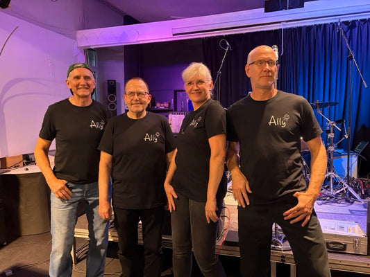 AllyBandMembers Four people standing together on a stage or backstage area, each wearing a black T-shirt with a white 'All' logo on the chest. The background shows stage equipment, cables, and curtains under purple and blue lighting.