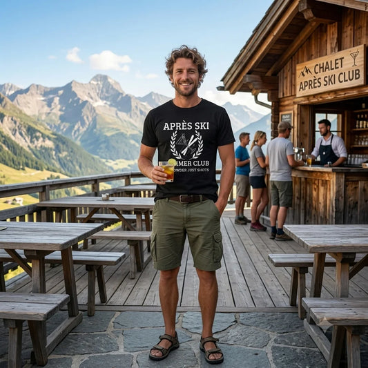Apres Ski Shirt A man stands on a mountain-side wooden chalet terrace wearing a black graphic T-shirt that reads “APRÈS SKI AFTER SKI CLUB” and olive green shorts, holding a drink. The background shows mountains, wooden tables, and a rustic lodge with people at the counter.