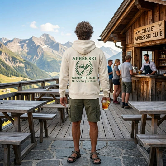 Après Ski Summer Club A man stands on a wooden outdoor patio at a mountain lodge, viewed from behind wearing a cream hoodie with a green 'APRÈS SKI SUMMER CLUB' graphic on the back. The background features a scenic mountain range, wooden picnic tables, and a rustic chalet with a sign reading 'CHALET APRÈS SKI CLUB'.