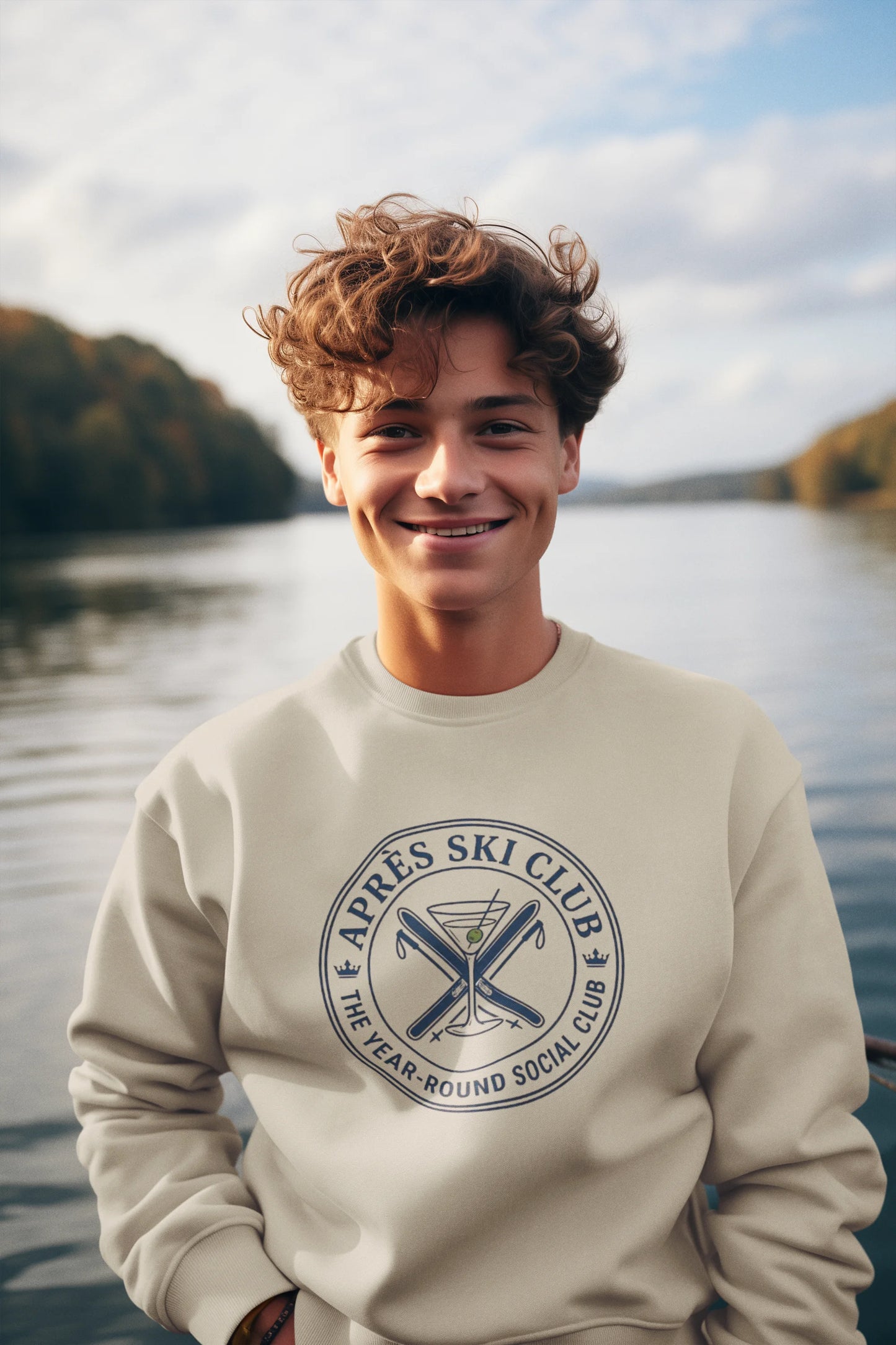 Après Ski Sweatshirt A smiling young man with curly brown hair stands outdoors by a lake wearing a cream sweatshirt with a dark blue circular graphic that reads 'Après Ski Club' and 'The Year Round Social Club.'.