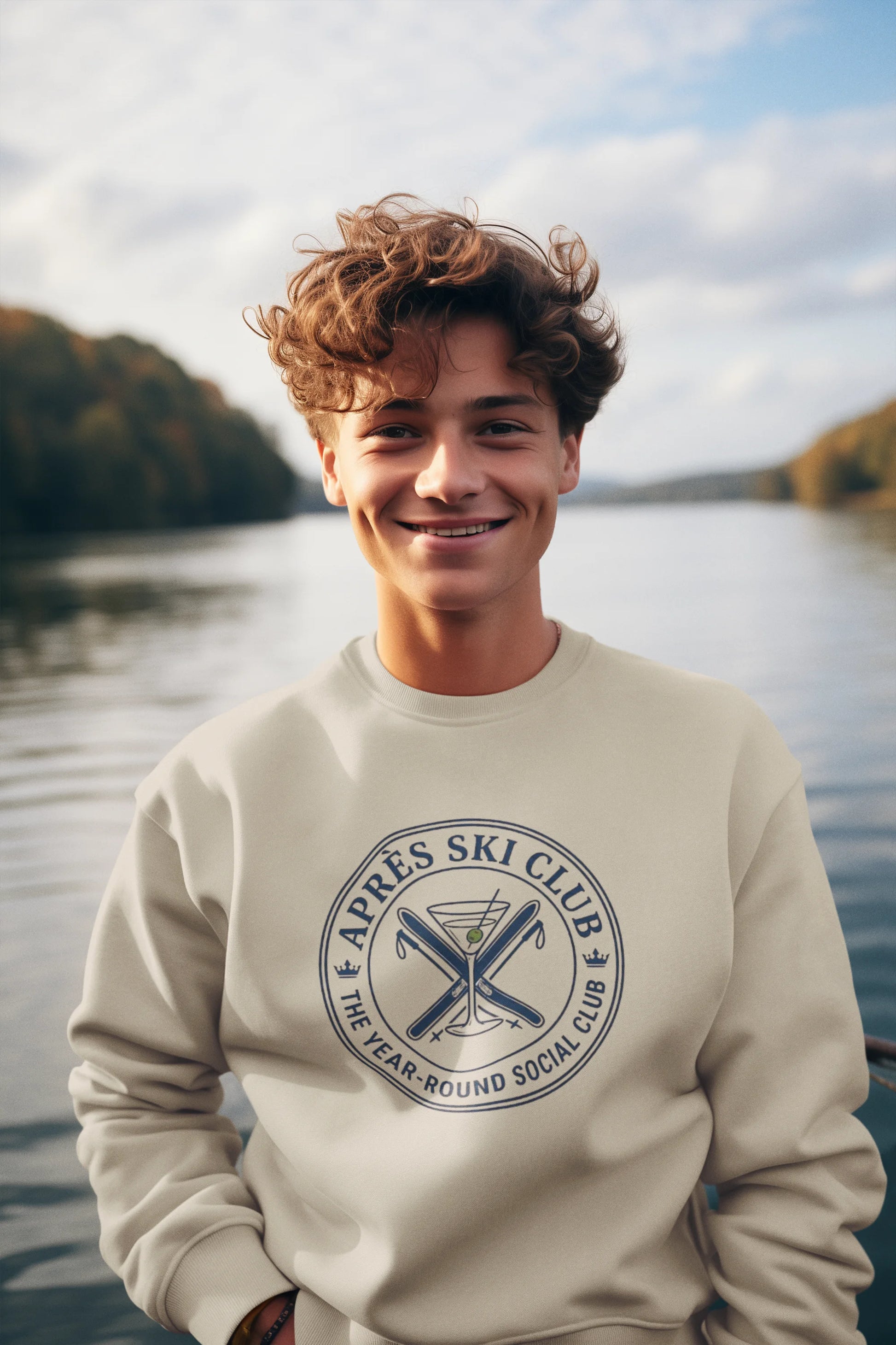 Après Ski Sweatshirt A smiling young man with curly brown hair stands outdoors by a lake wearing a cream sweatshirt with a dark blue circular graphic that reads 'Après Ski Club' and 'The Year Round Social Club.'.
