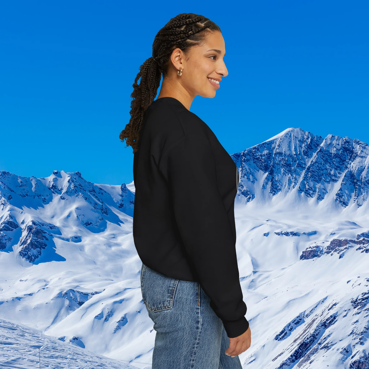 Apres Ski sweater A woman with braided hair is shown in profile wearing a black long-sleeve top and light blue jeans, set against a bright blue sky and snowy mountain backdrop.