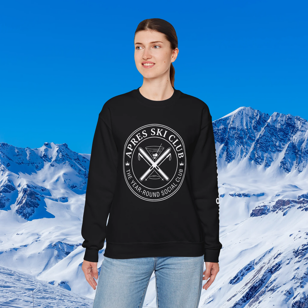 Apres Ski sweater A woman modeling a black crewneck sweatshirt with a white circular graphic that reads 'APRES SKI CLUB THE YEAR-ROUND SOCIAL CLUB' over crossed skis, set against a snowy mountain and blue sky background.