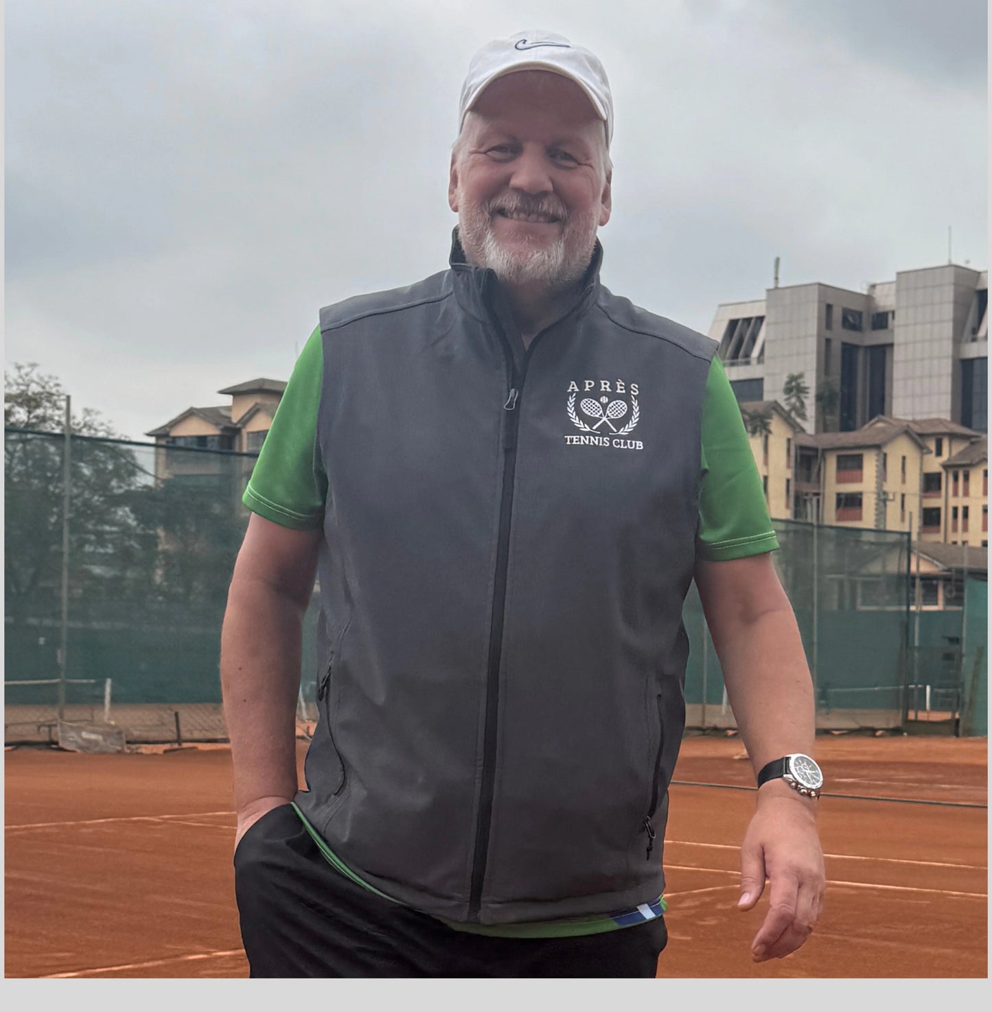 Apres Tennis Vest A smiling man wearing a white cap, a green short-sleeve shirt, and a dark gray zip-up athletic vest with a white 'APPEX TENNIS CLUB' logo on the chest, standing on an outdoor clay tennis court.