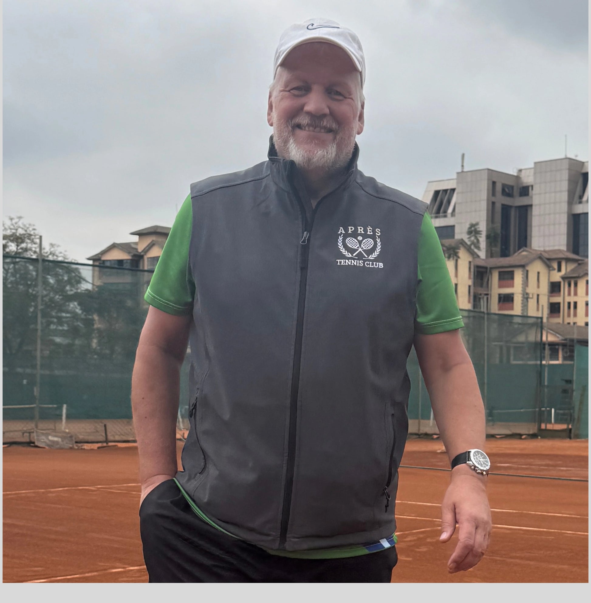 Apres Tennis Vest A smiling man wearing a white cap, a green short-sleeve shirt, and a dark gray zip-up athletic vest with a white 'APPEX TENNIS CLUB' logo on the chest, standing on an outdoor clay tennis court.