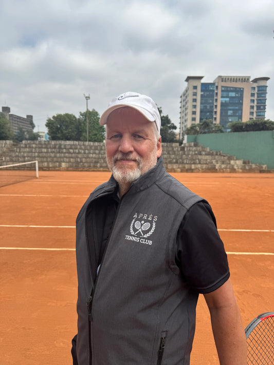 Apres Tennis Vest A man wearing a dark gray zip-up vest with a white tennis logo and a white cap, standing on an outdoor clay tennis court with stadium seating and buildings in the background.