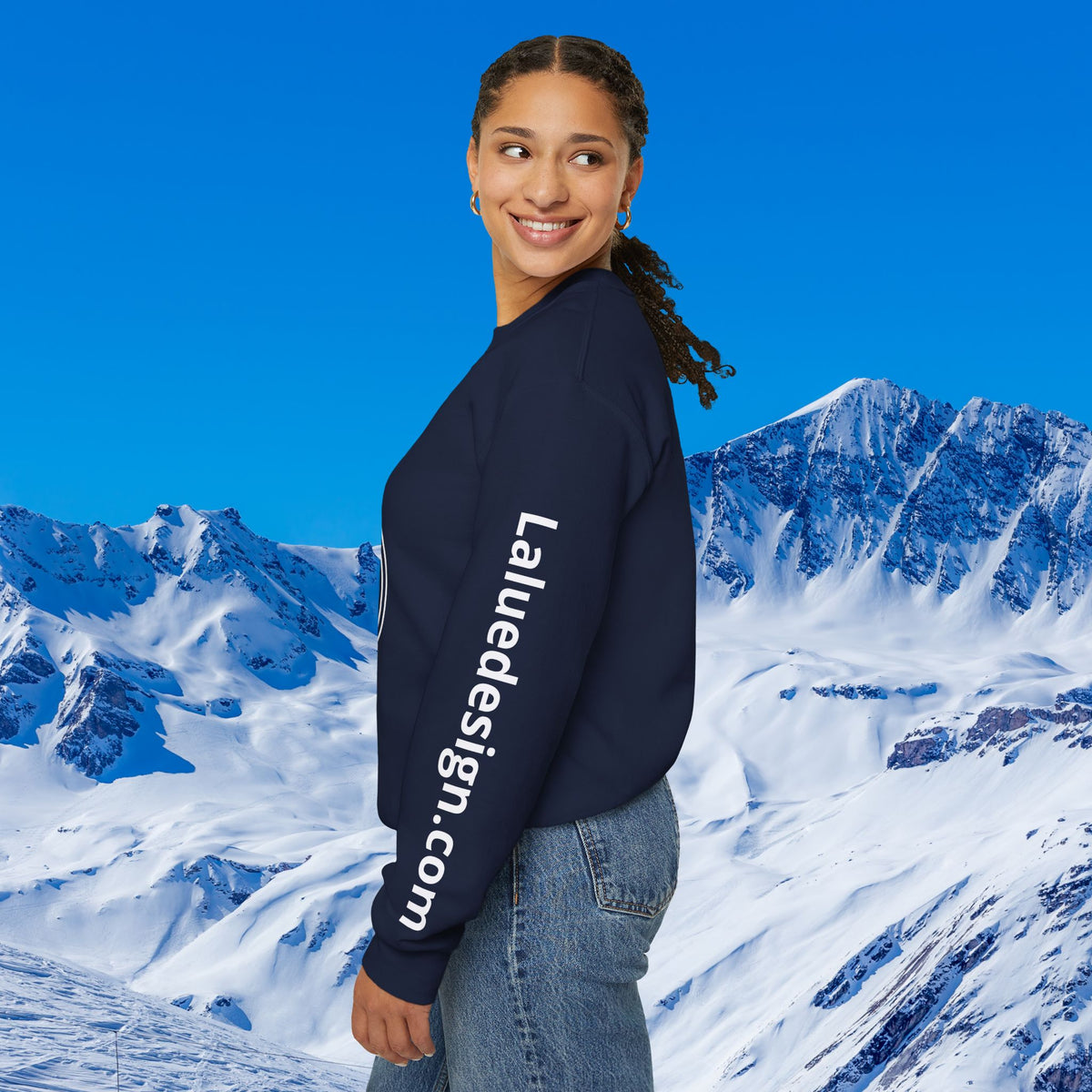 Apres Ski sweater A woman wearing a navy blue crewneck sweatshirt with 'LaudeDesign.com' printed in white down the left sleeve, posed against a backdrop of snowy mountains and a clear blue sky.