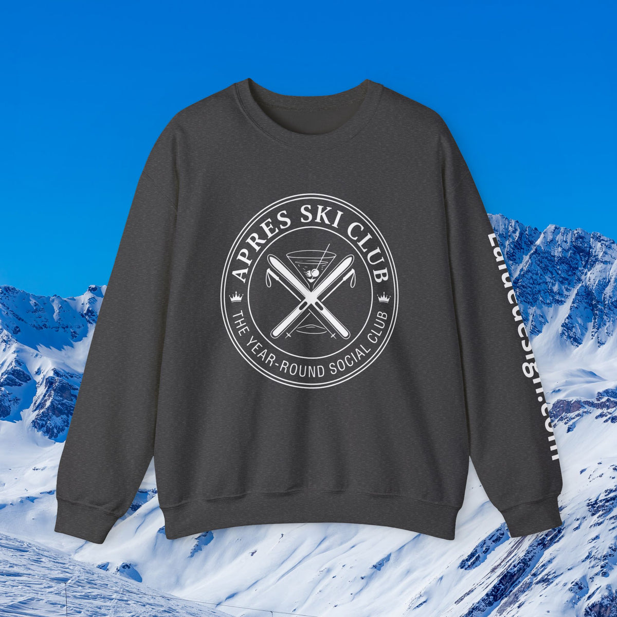 Apres Ski sweater A dark gray crewneck sweatshirt with a white circular graphic reading 'APRES SKI CLUB' and 'THE YEAR-ROUND SOCIAL CLUB' over crossed skis, shown against a snowy mountain background under a blue sky. White text is printed down the right sleeve.