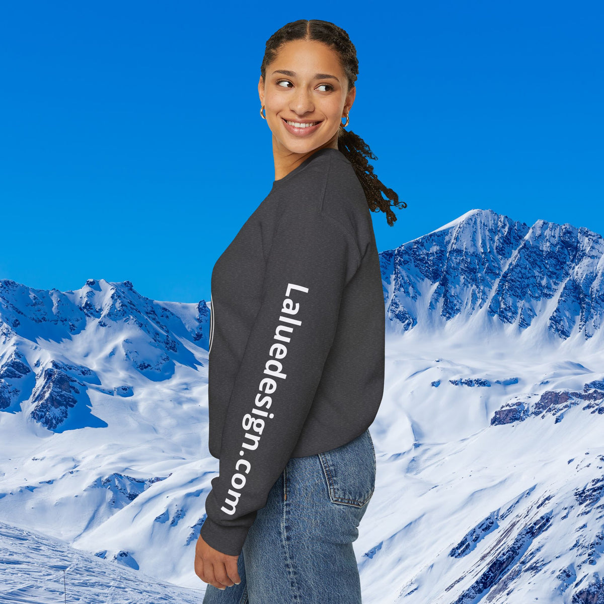Apres Ski sweater A woman wearing a dark gray crewneck sweatshirt with 'LaudeDesign.com' printed in white down the left sleeve, posed against a digital background of snowy mountains and a blue sky.