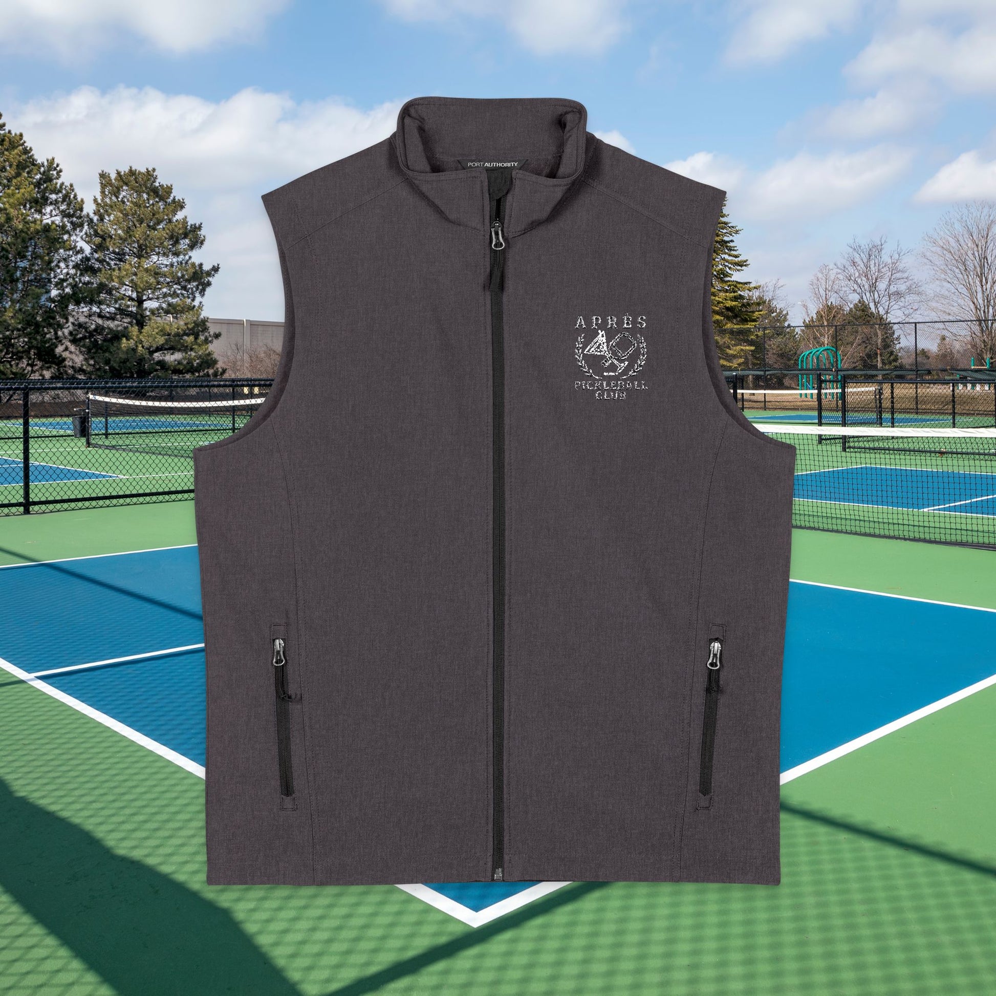 Apres pickleball vest A dark gray sleeveless zip-front vest with a stand collar and two vertical zip pockets, shown centered over a tennis court background. The left chest has a white printed crest/logo reading ARBES UNIVERSITY CARE.