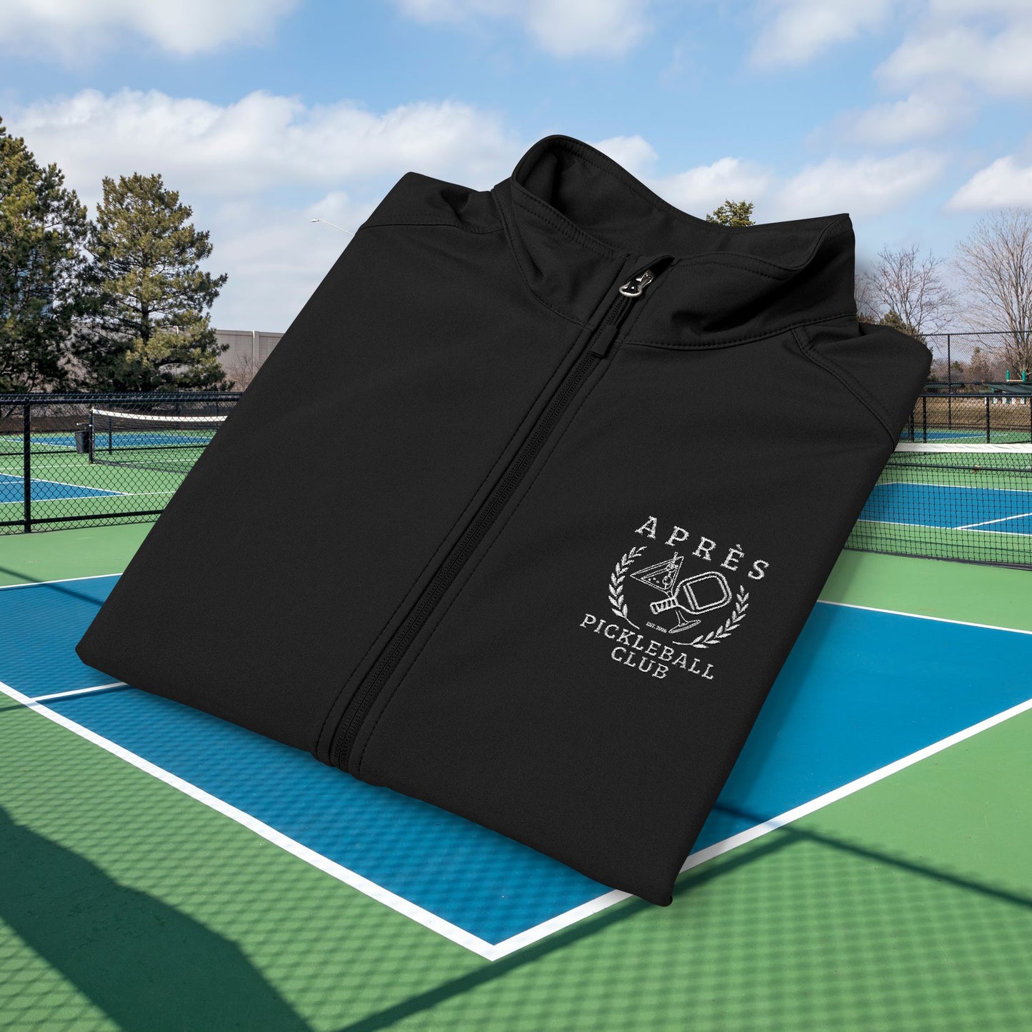 Apres pickleball vest A black zip-up jacket with a white 'Après Pigeûball Club' logo on the chest, displayed on a green and blue outdoor pickleball court under a blue sky.