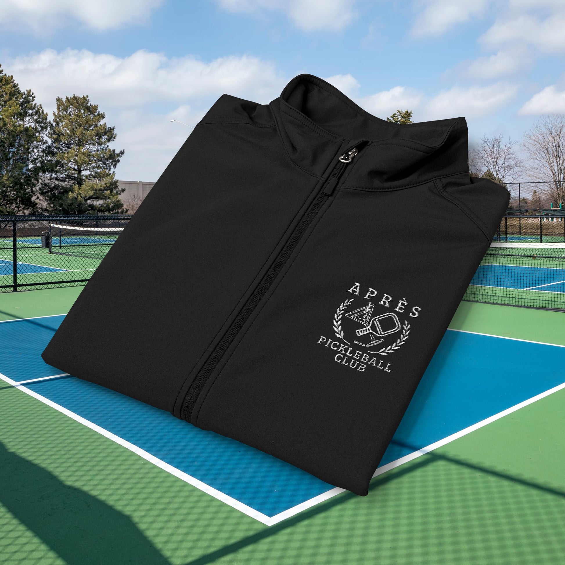 Apres pickleball vest A black zip-up jacket with a white 'Après Pigeûball Club' logo on the chest, displayed on a green and blue outdoor pickleball court under a blue sky.