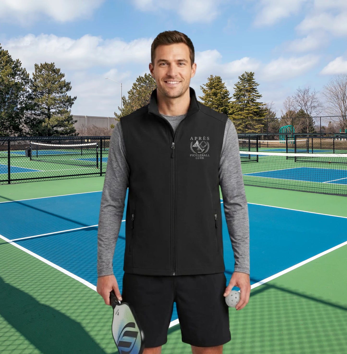 Apres pickleball vest A man stands on a blue and green tennis court wearing a black zip-up vest over a gray long-sleeve shirt and black shorts, holding a pickleball paddle and ball. The background shows other tennis courts, trees, and a blue sky with clouds.