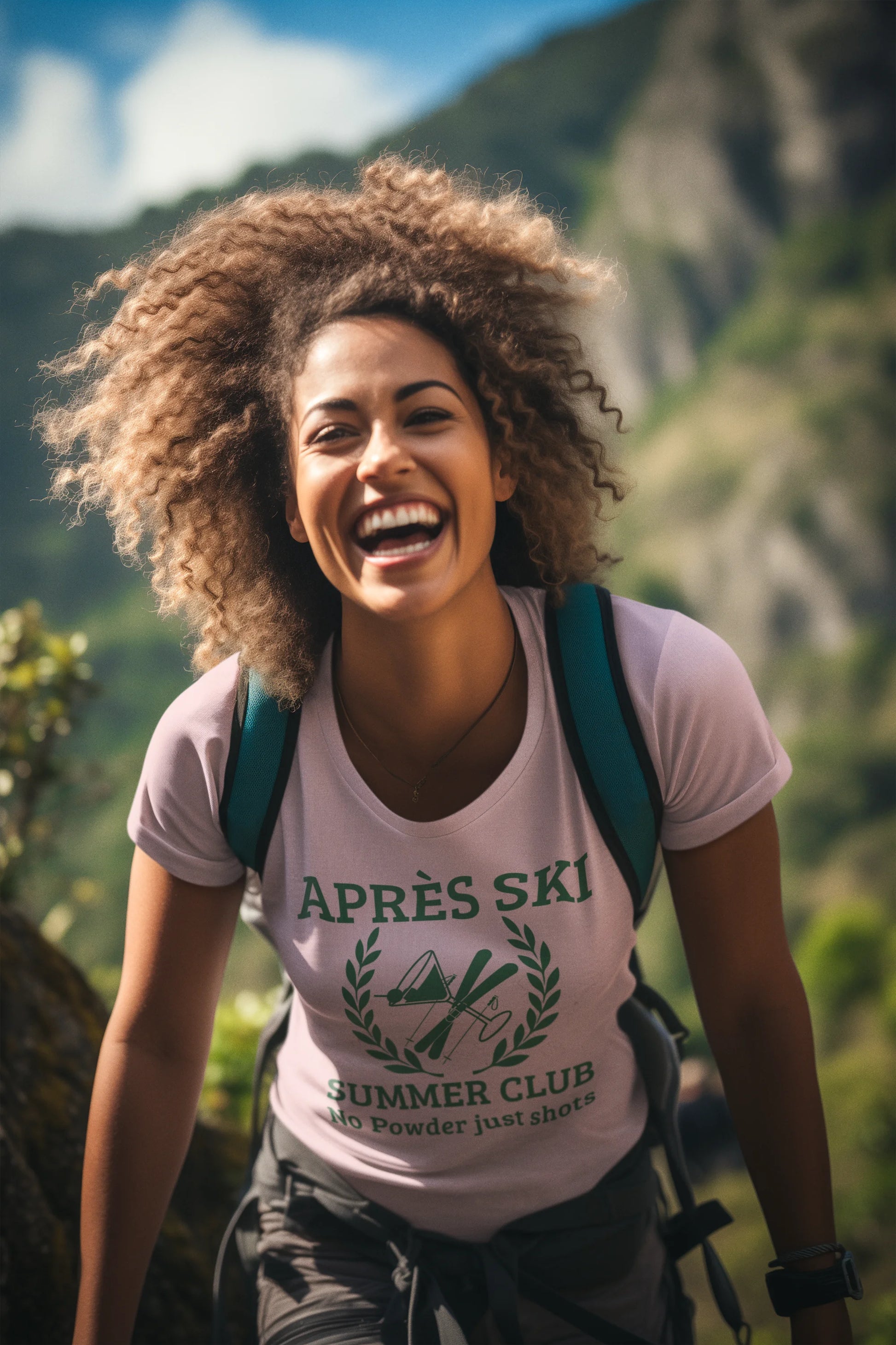 Apres ski clothes ladies A smiling woman outdoors in front of mountains, wearing a light pink graphic T-shirt that reads 'APRÈS SKI SUMMER CLUB' with a green graphic print and a dark green backpack.