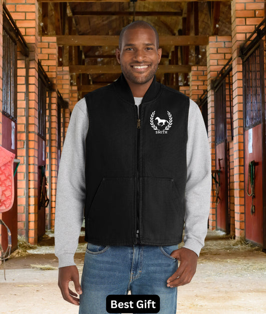 Equestrian vest A man wearing a black zip-up vest with gray sleeves and a white horse logo on the chest, standing in a stable corridor. A black label at the bottom reads 'Best Gift'.