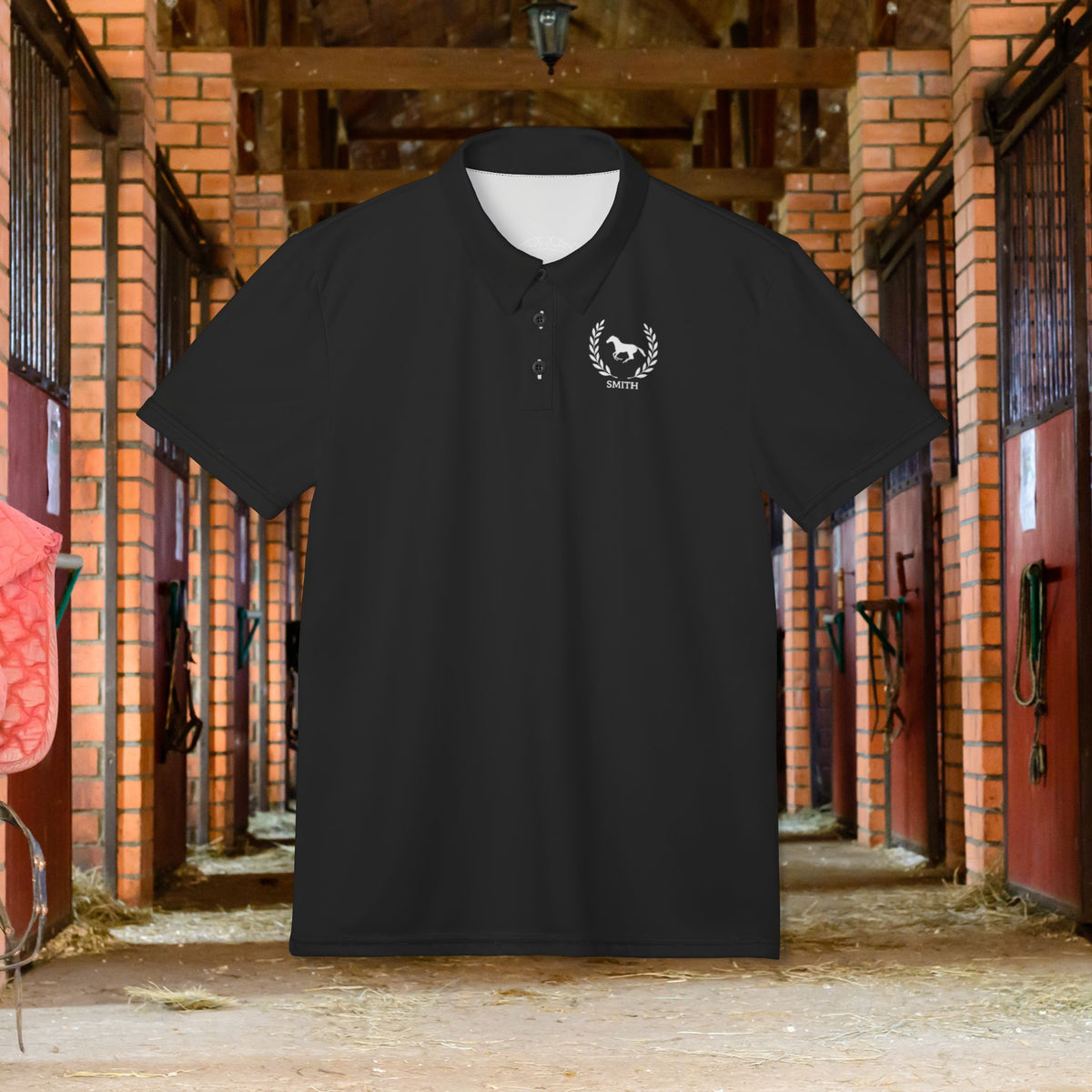 Equestrian polo shirt A black short-sleeve polo shirt with a small white horse-and-lawn crest logo on the left chest, displayed against a blurred background of an equestrian stable interior.