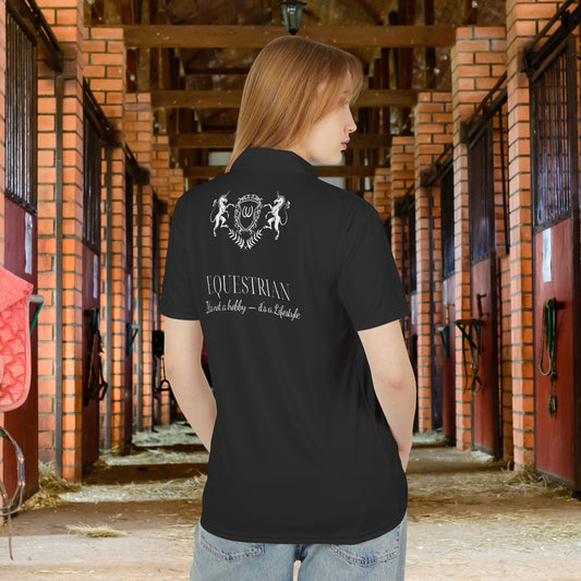 Equestrian polo shirt A woman standing in a stable corridor wearing a black polo shirt with a white equestrian crest and 'EQUESTRIAN' text on the back, paired with blue jeans.