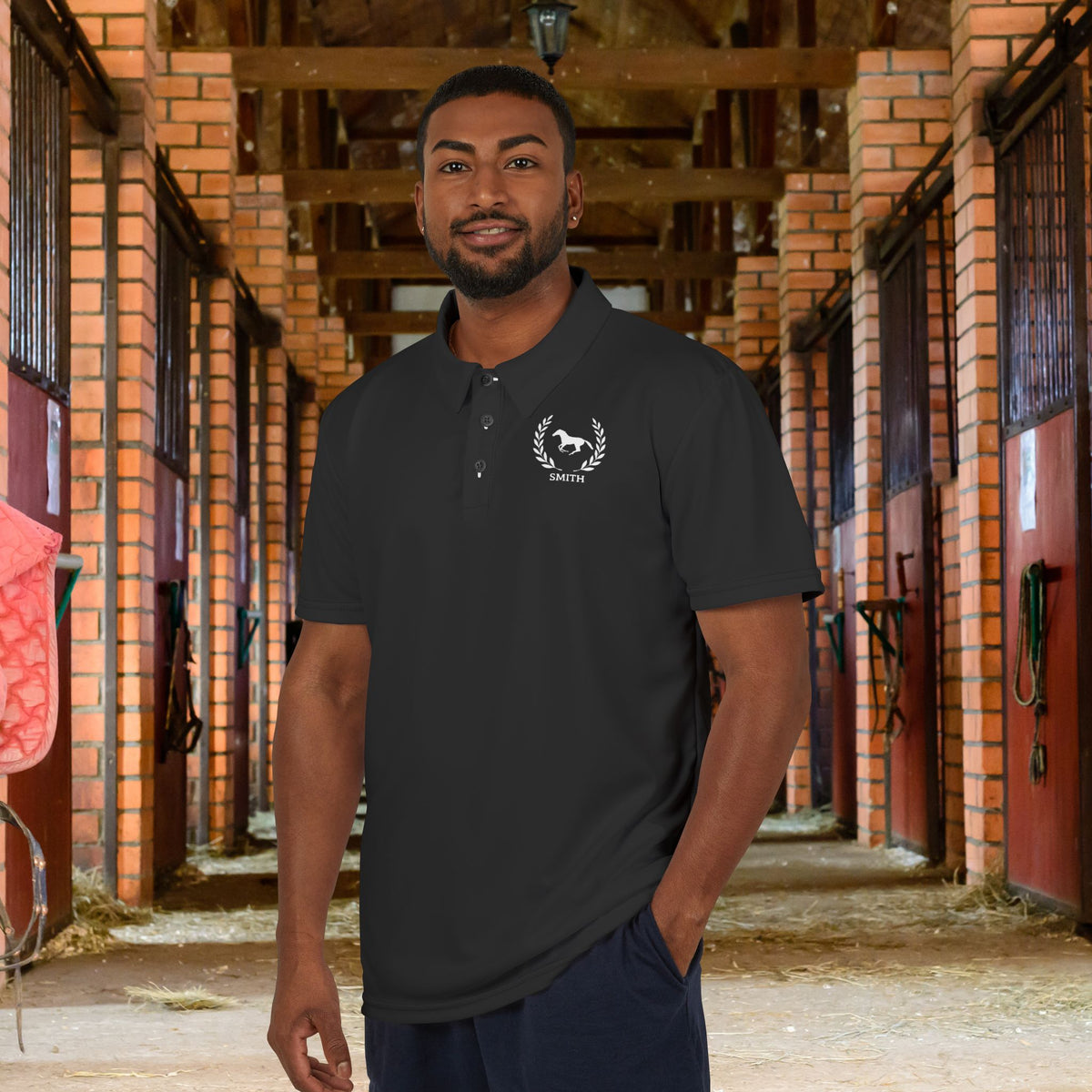 Equestrian polo shirt A man wearing a dark charcoal gray polo shirt with a white horse logo and the word 'STOCKTON' on the chest, standing in a brick horse stable corridor.