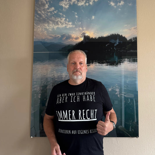 A man wearing a black graphic t-shirt with white German text stands next to a large vertical wall art print depicting a scenic lake reflection under a cloudy sky. The photo is taken from above against a beige wall.