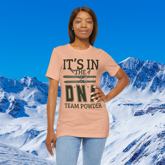 It's in the DNA A woman modeling a heather peach t-shirt with a dark green graphic that reads 'IT'S IN THE DNF TEAM POWDER' against a snowy mountain and blue sky background.
