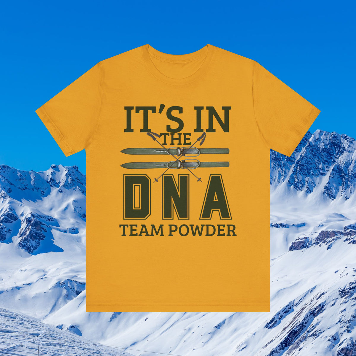 It's in the DNA A yellow graphic t-shirt with the text 'IT'S IN THE DNA TEAM POWDER' and skis printed on the front, set against a snowy mountain background under a blue sky.