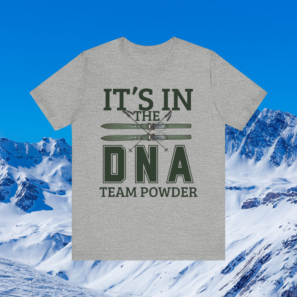 It's in the DNA A heather gray short-sleeve t-shirt with a dark green graphic reading 'IT'S IN THE DNA TEAM POWDER' and crossed skis, shown against a snowy mountain and blue sky background.