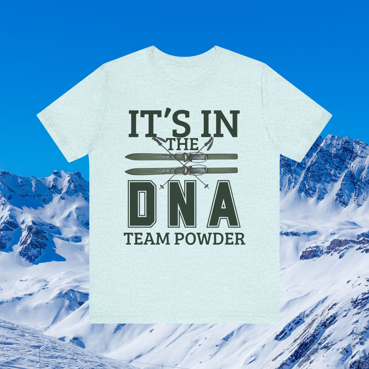 It's in the DNA A light heather blue short-sleeve t-shirt with a dark green graphic reading 'IT'S IN THE DNA TEAM POWDER' and crossed skis, shown against a snowy mountain and blue sky background.
