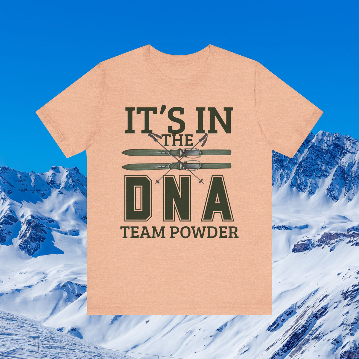 It's in the DNA A heather peach short-sleeve t-shirt with a dark green graphic reading 'IT'S IN THE DNA TEAM POWDER' and crossed skis, shown against a snowy mountain and blue sky background.