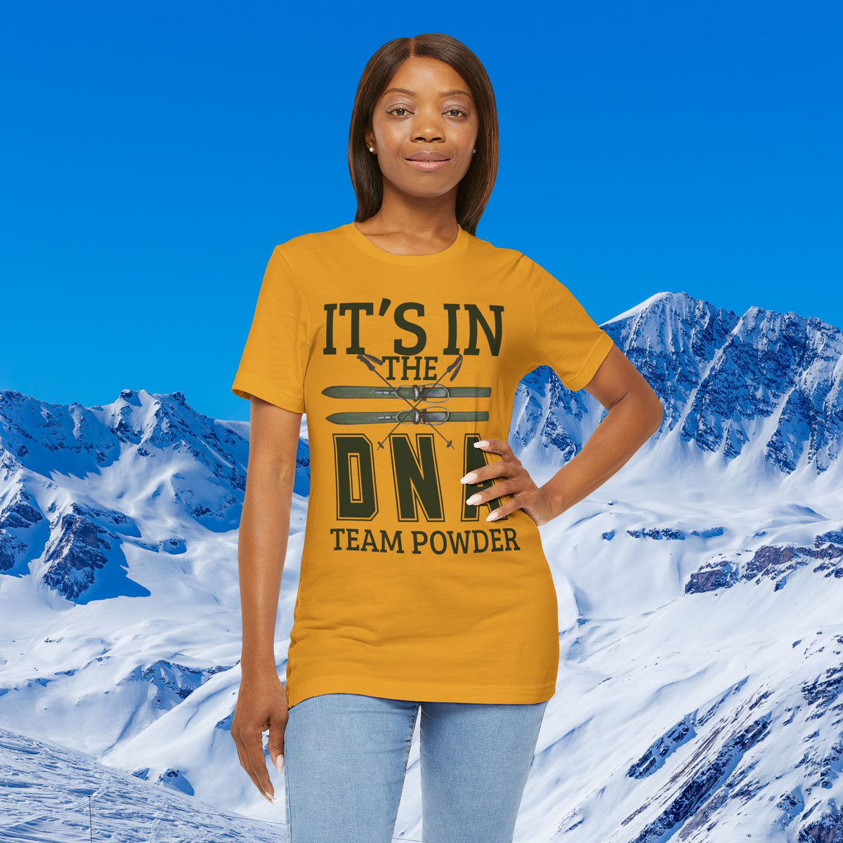 It's in the DNA A woman modeling a mustard yellow graphic t-shirt that reads 'IT'S IN THE DNF TEAM POWDER' with a double-hockey stick design, set against a snowy mountain and blue sky background.