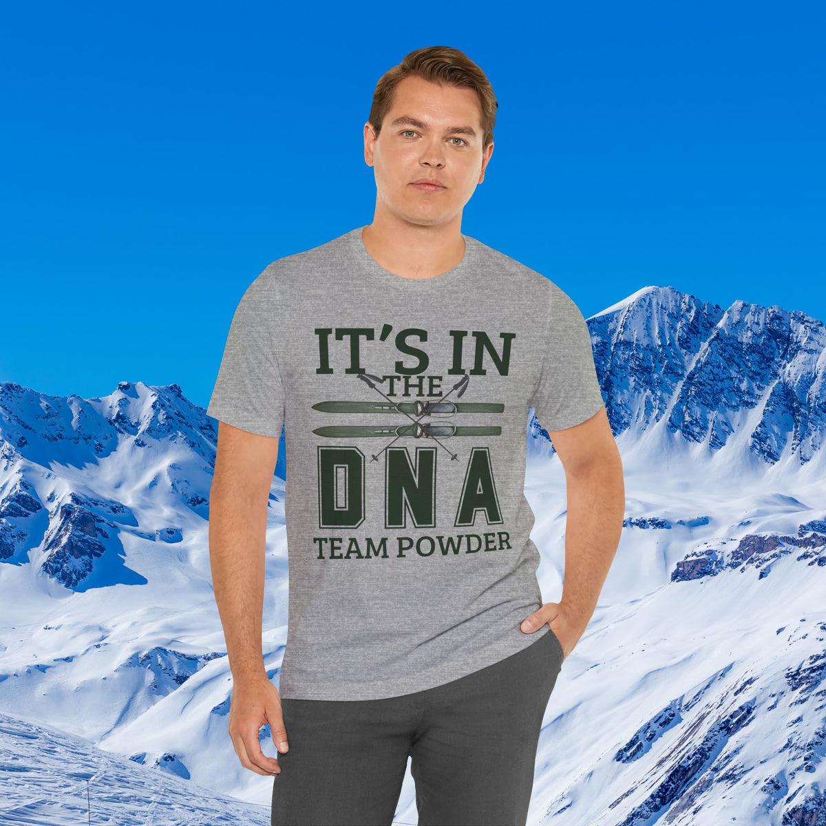 It's in the DNA A man wearing a heather gray graphic t-shirt with the text 'IT'S IN THE DNA TEAM POWDER' and a small airplane graphic, posed against a digital background of snowy mountains and a blue sky.