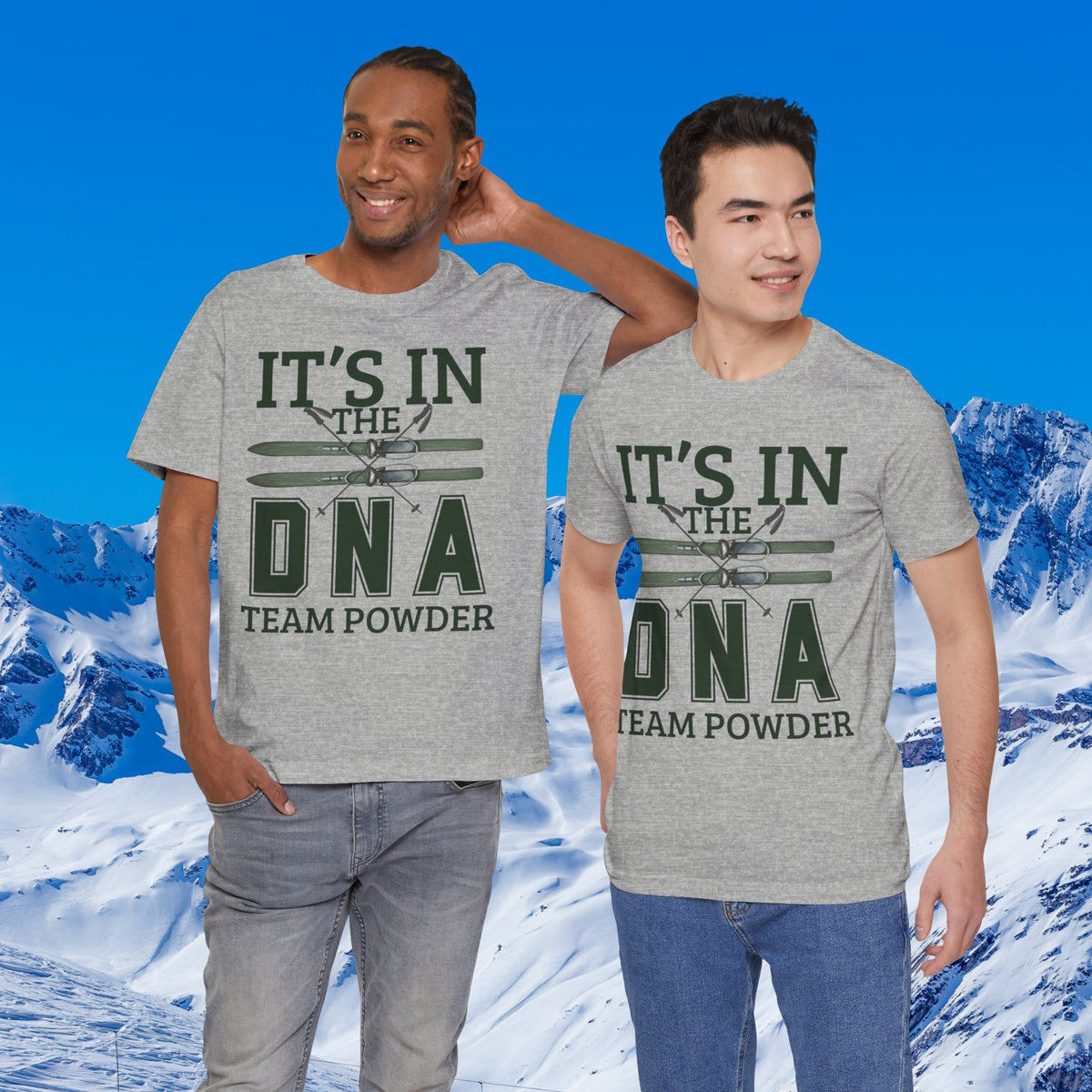 It's in the DNA Two men modeling matching heather gray graphic t-shirts with a dark green and white 'IT'S IN THE DNA TEAM POWDER' design, set against a snowy mountain background under a blue sky.