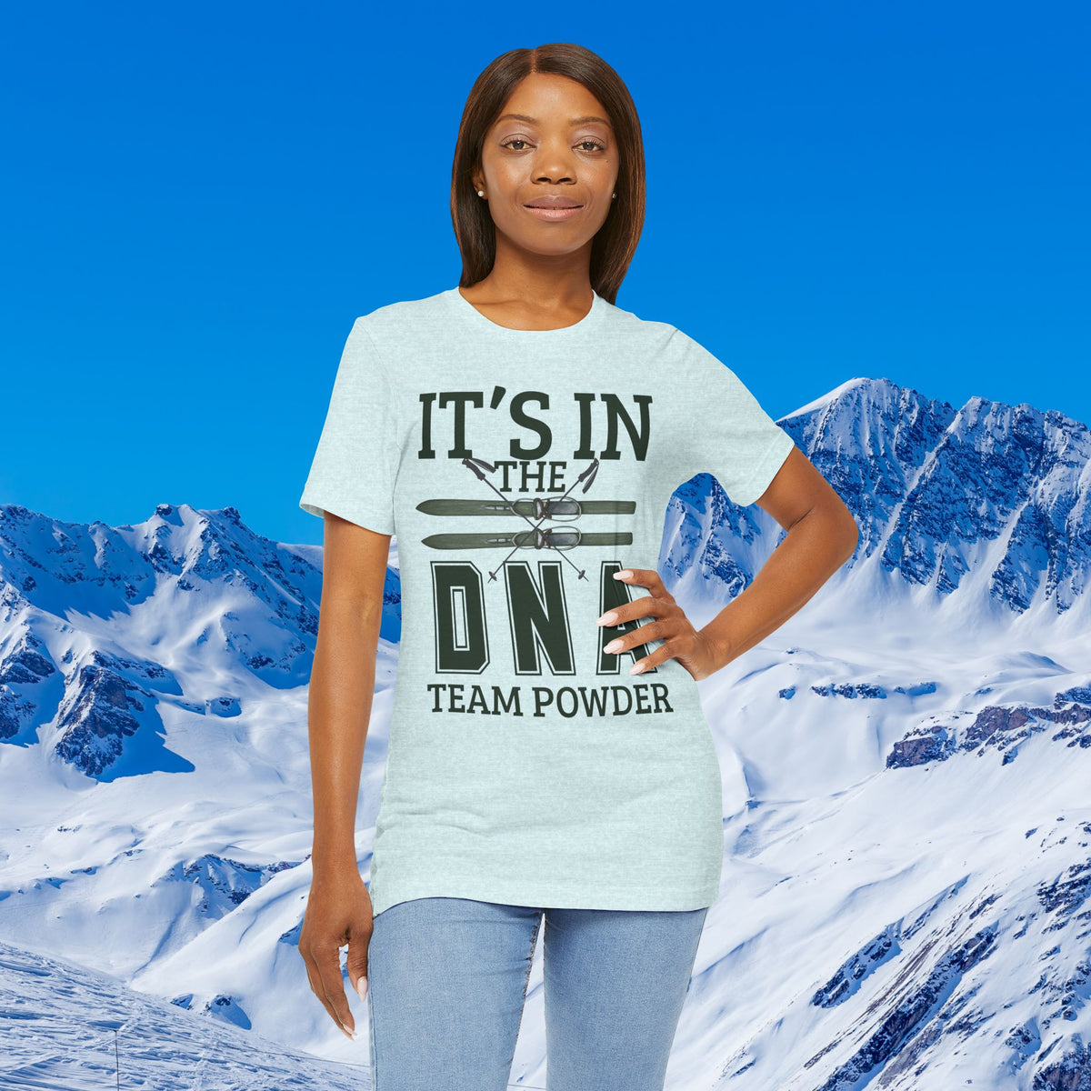 It's in the DNA A woman modeling a light heather gray graphic t-shirt with the text 'IT'S IN THE DNF TEAM POWDER' and a ski graphic, set against a snowy mountain and blue sky background.