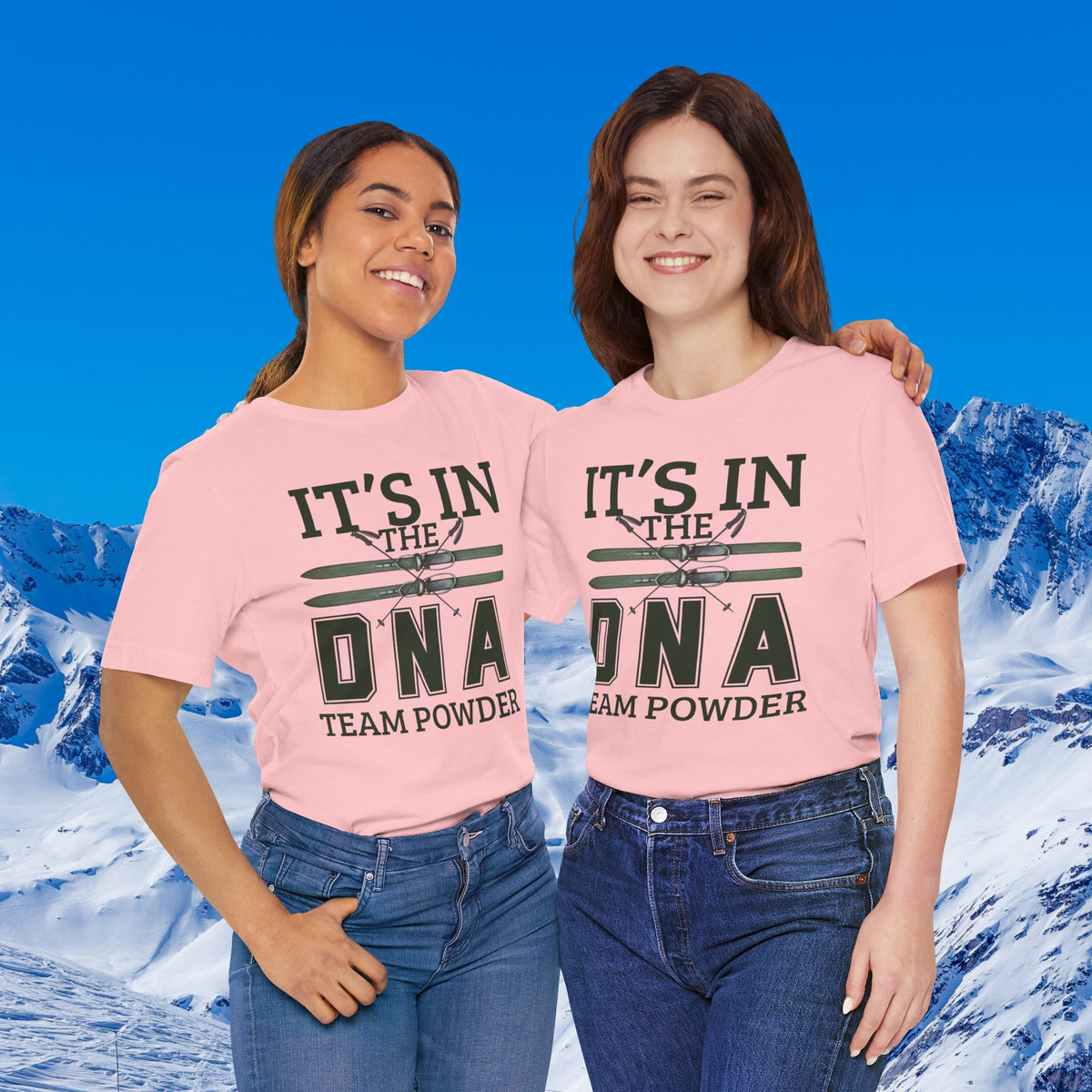 It's in the DNA Two women standing together in front of a snowy mountain backdrop, wearing matching light pink graphic T-shirts with dark green and black text that reads 'IT'S IN THE DNA TEAM POWDER.'.