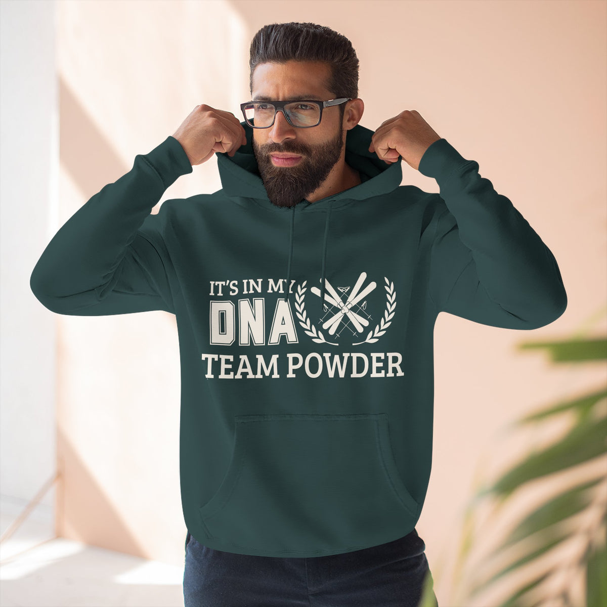 Its in my DNA A man wearing a dark green hoodie with a white graphic print that reads 'IT'S IN MY DNA TEAM POWDER' and flexing his arms. The background is a soft beige wall with a blurred plant in the foreground.