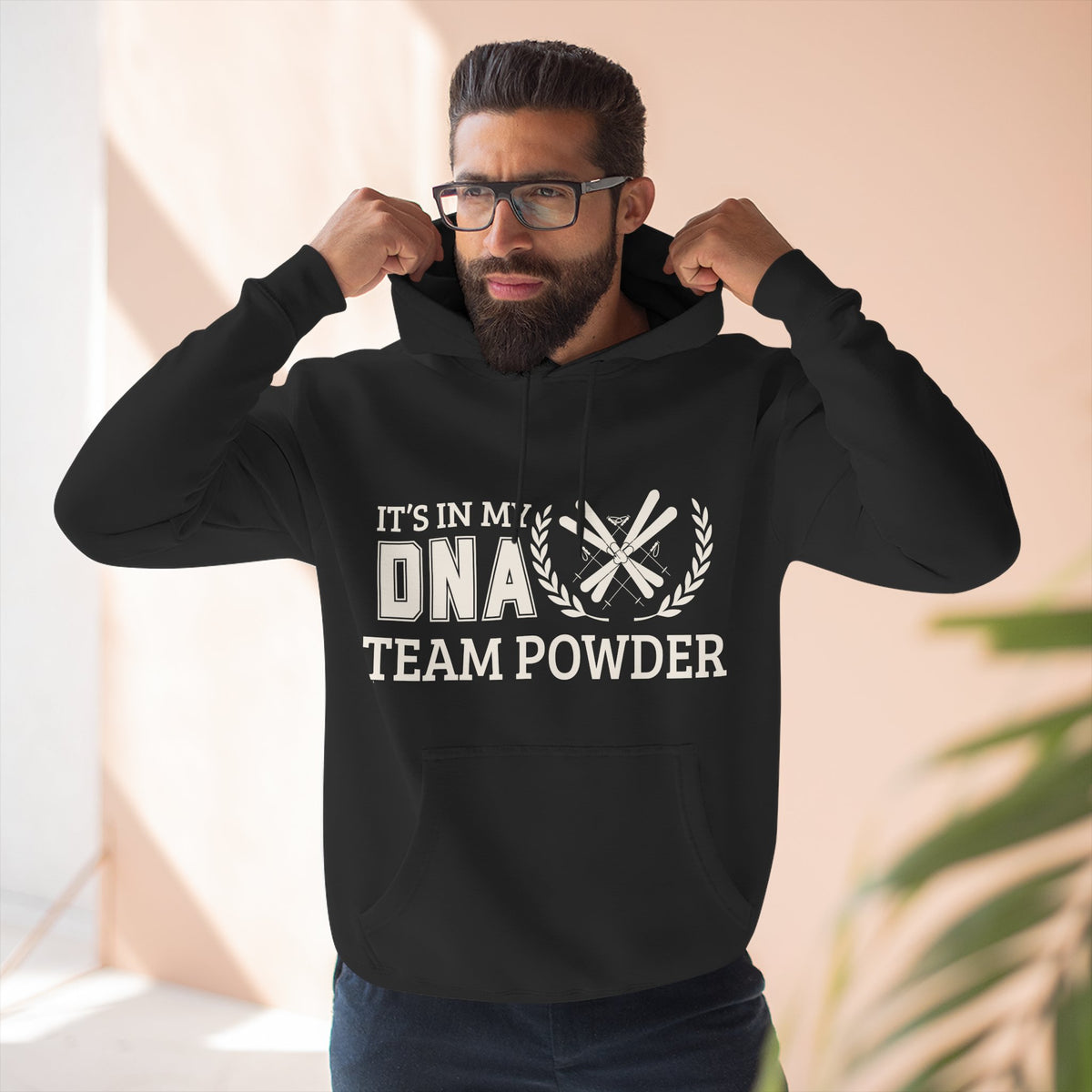Its in my DNA A man wearing a black hoodie with a white graphic print that reads 'IT'S IN MY DNA TEAM POWDER' and 'DNA' replaced by crossed bones. He is posing with his hands on the hood of the sweatshirt in a brightly lit indoor setting.
