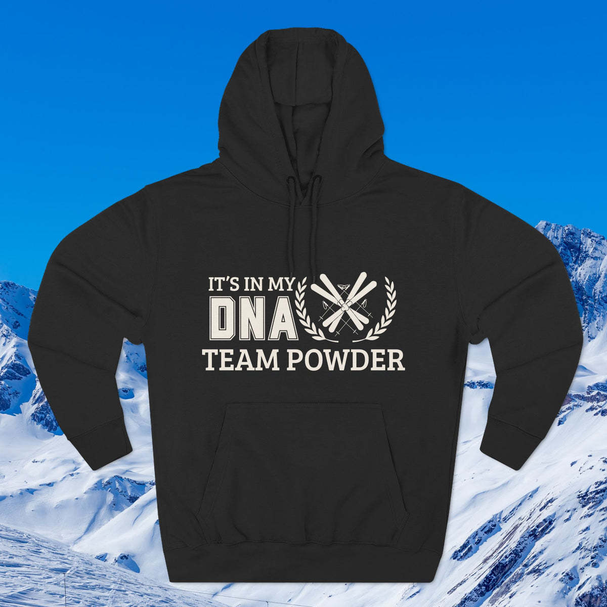 Its in my DNA A black pullover hoodie with a distressed white graphic reading 'IT'S IN MY DNA TEAM POWDER' and crossed ski poles with laurel branches, shown against a blue sky and snowy mountain backdrop.