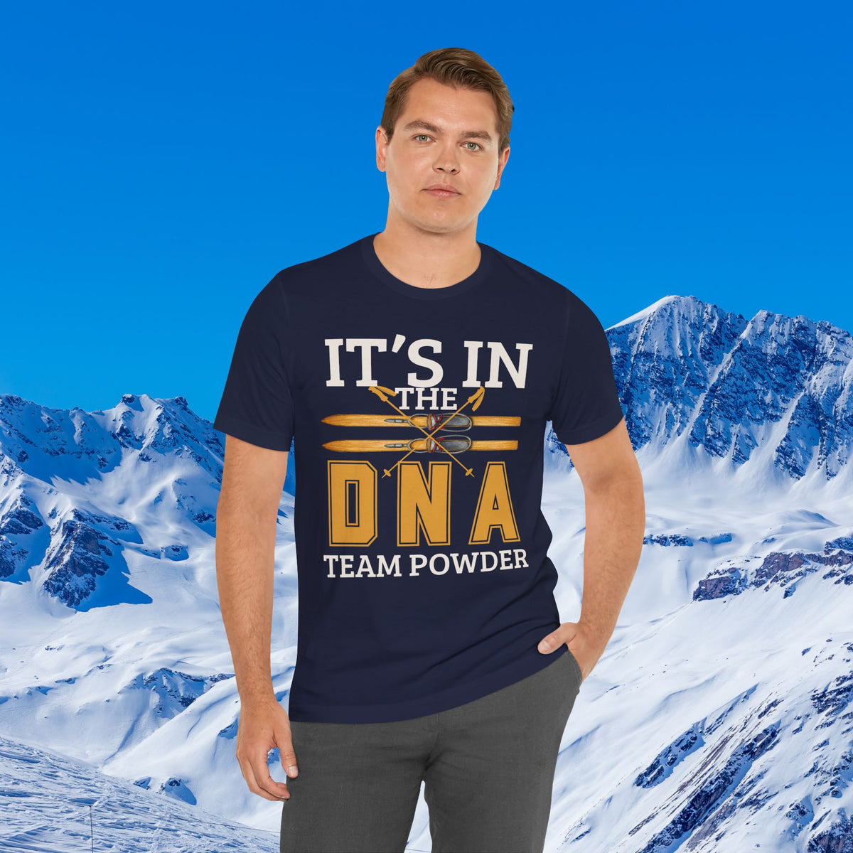 Its in the DNA A man wearing a navy blue graphic t-shirt with the text 'IT'S IN THE DNA TEAM POWDER' in white and gold, posed against a snowy mountain and blue sky background.