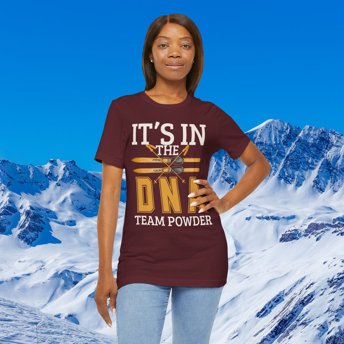 Its in the DNA A woman modeling a maroon short-sleeve graphic T-shirt with the text 'IT'S IN THE DND TEAM POWDER' in white and gold, set against a snowy mountain and blue sky background.