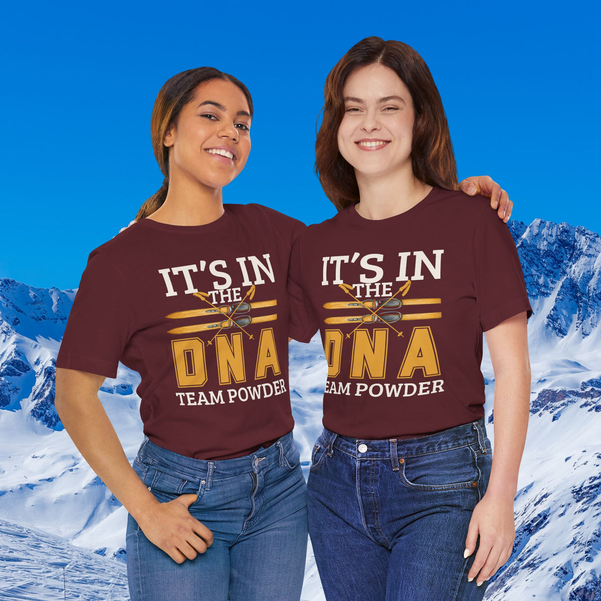 Its in the DNA Two women standing side by side in front of a snowy mountain backdrop, wearing matching maroon graphic T-shirts with the text 'IT'S IN THE DNA TEAM POWDER' in white and gold lettering.